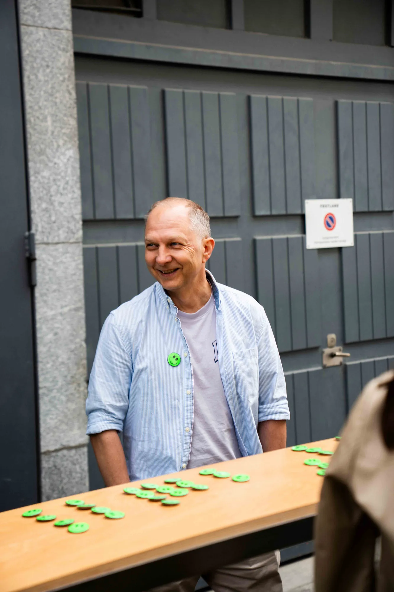 Lächelnder Mann im hellblauen Hemd steht hinter einem Holztisch mit ausgelegten grünen Smiley-Buttons vor einem grauen Holztor.