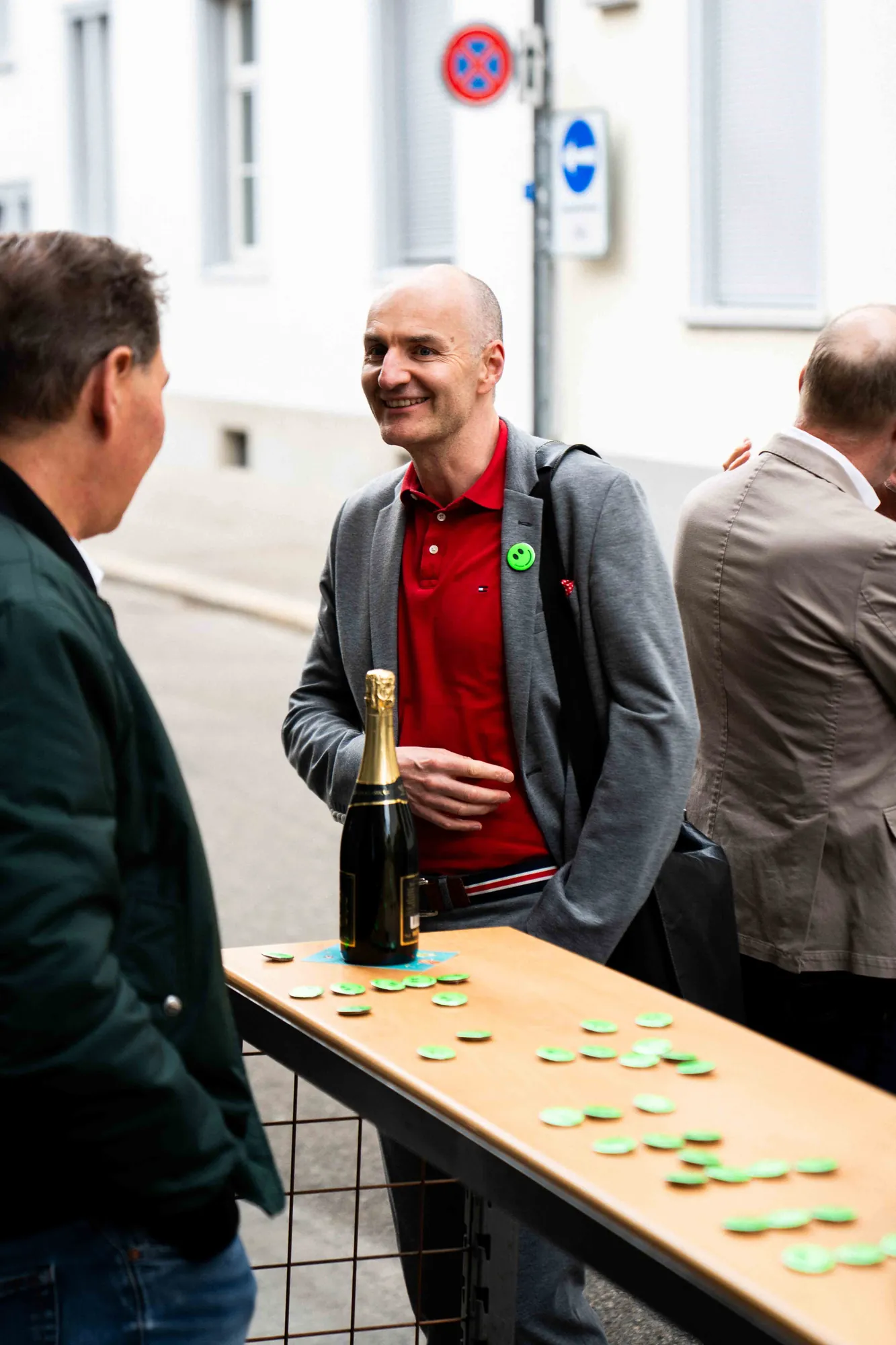 Gast im roten Poloshirt unterhält sich draussen an einem Stehtisch mit Champagnerflasche und verstreuten grünen Smiley-Buttons.