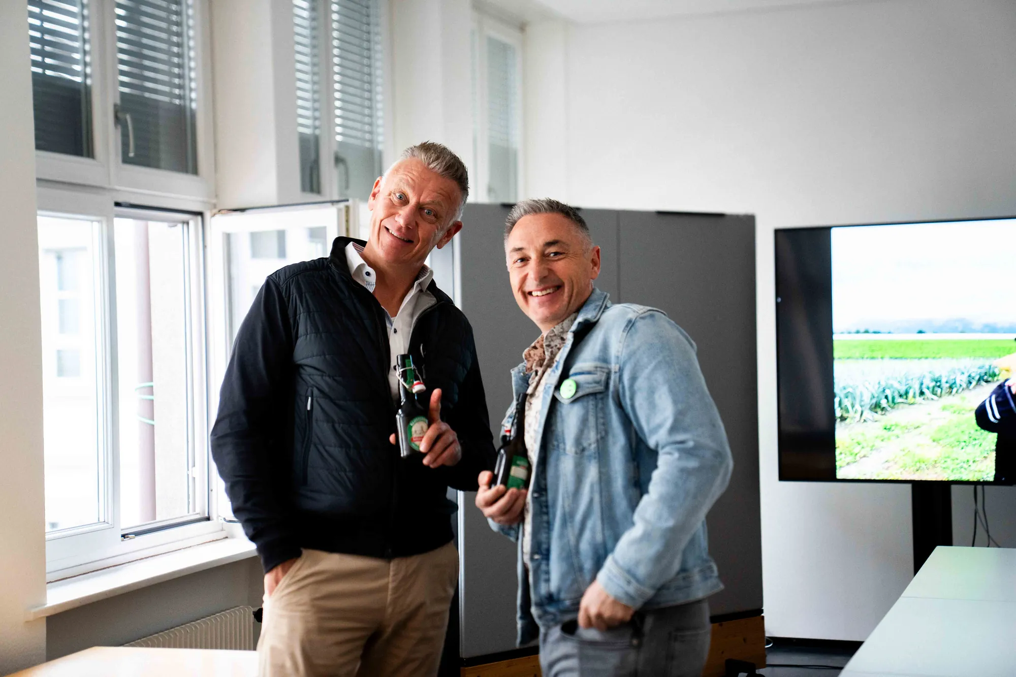 Zwei Männer posieren gut gelaunt mit Bierflaschen im Büro, rechts ein Bildschirm mit Foto eines Gemüsefelds.