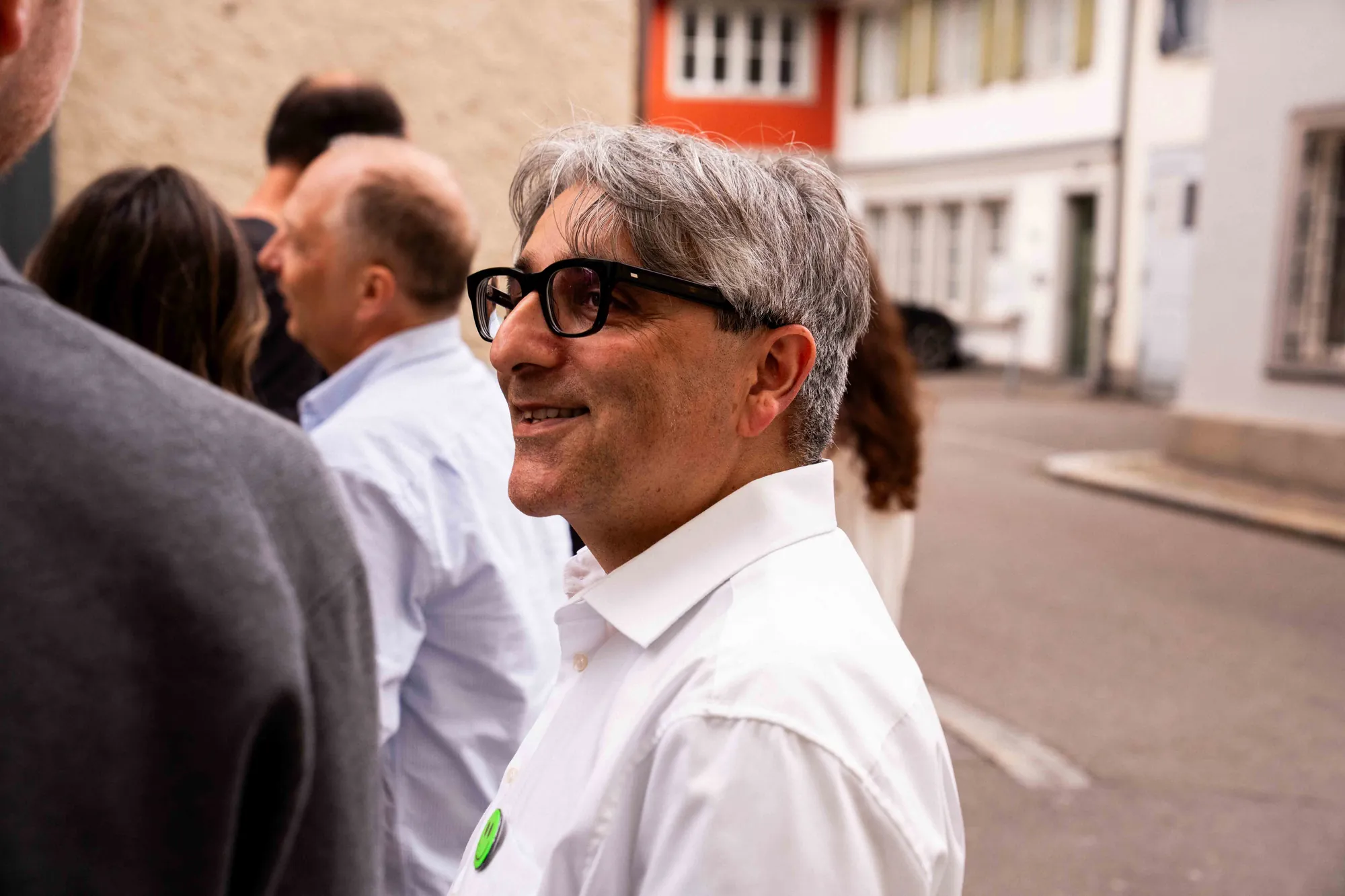 Mann mit dunkler Hornbrille und weissem Hemd blickt lächelnd zur Seite, um ihn herum Gäste auf der Gasse vor der Agentur.
