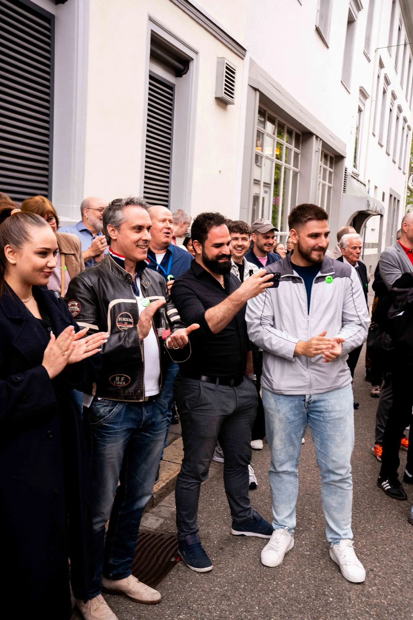 Publikum steht dicht gedrängt vor einer weissen Hausfassade auf der Strasse, applaudiert und filmt das Geschehen mit dem Handy.