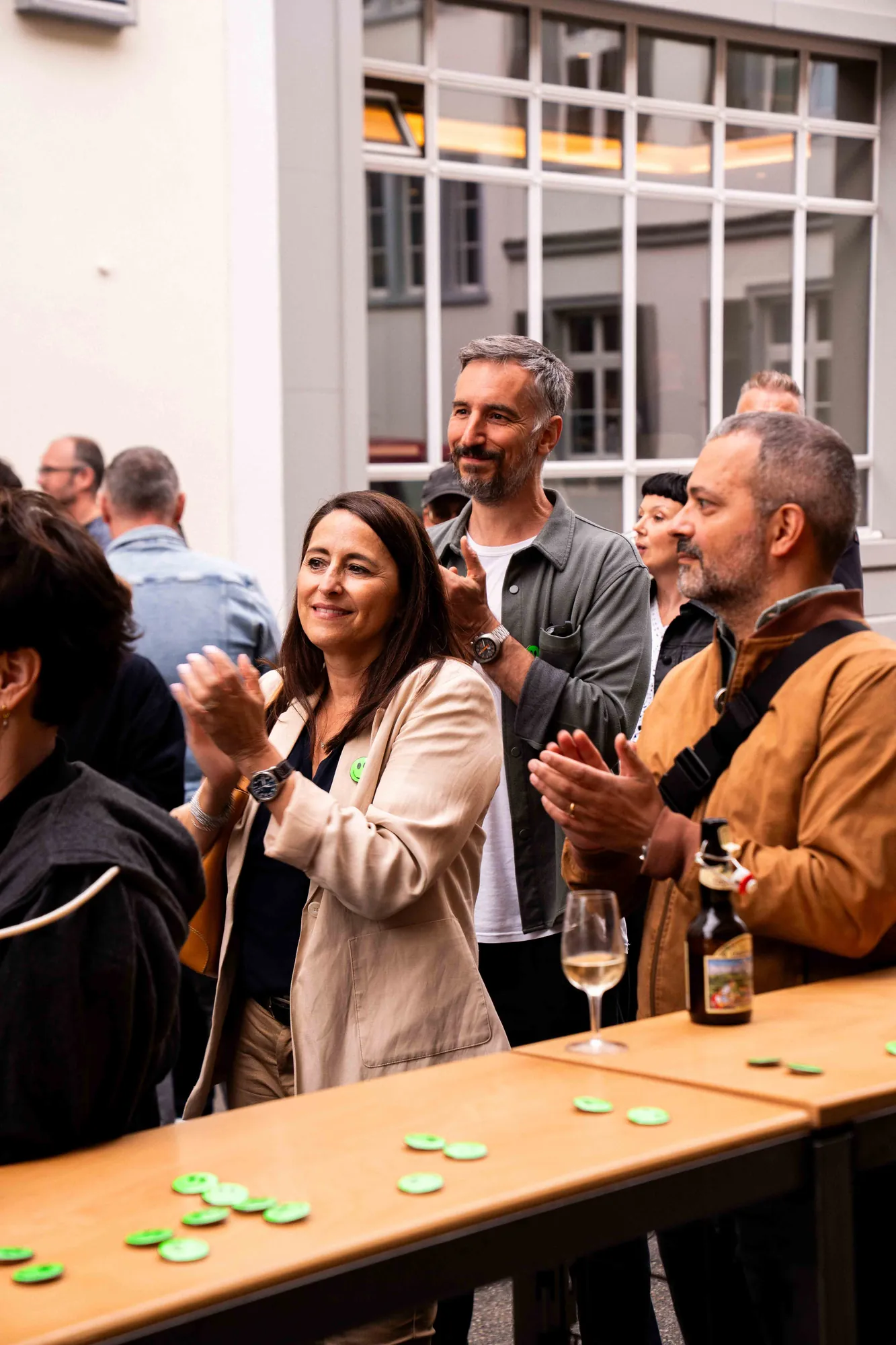 Lächelnde Gäste applaudieren an einem Stehtisch mit Weinglas, Bierflasche und grünen Buttons vor einer grossen Fensterfront.