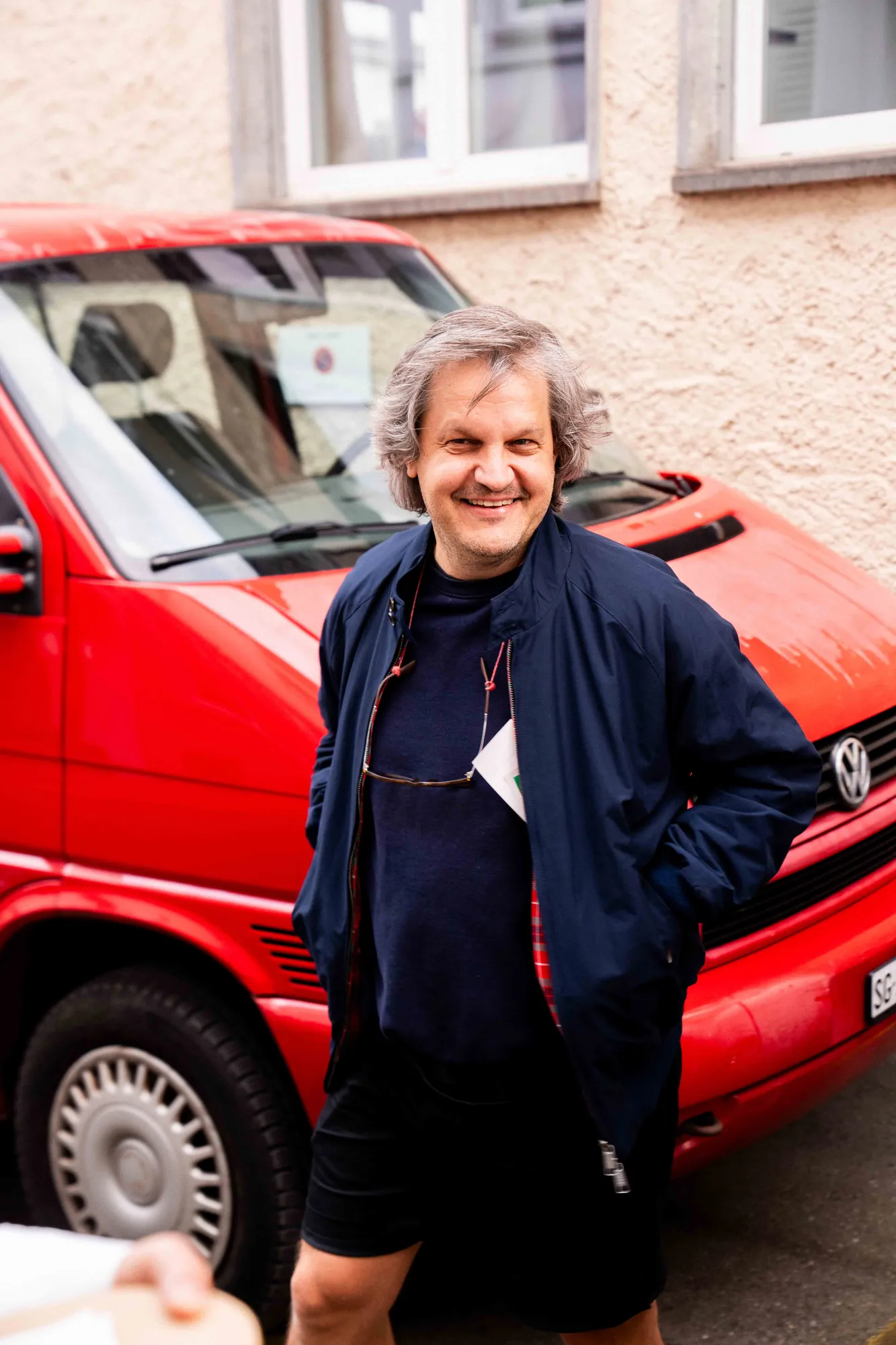 Lächelnder Mann in dunkelblauer Jacke steht mit den Händen in den Taschen vor einem roten VW-Bus in einer Gasse.