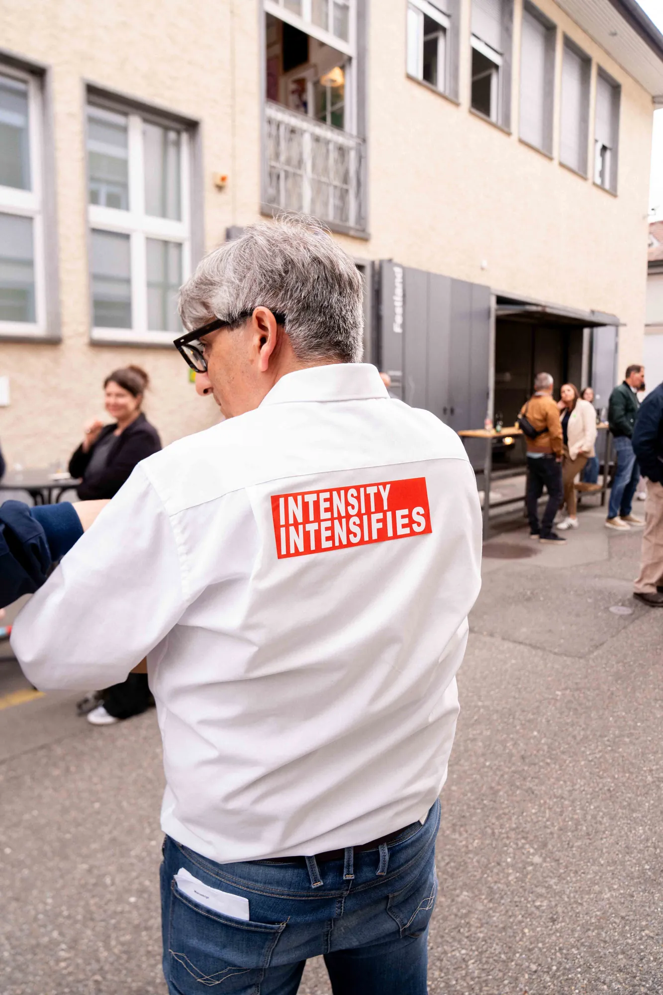 Rückenansicht eines Mannes im weissen Hemd mit rotem Aufnäher «Intensity Intensifies», im Hintergrund plaudernde Gäste im Hof.