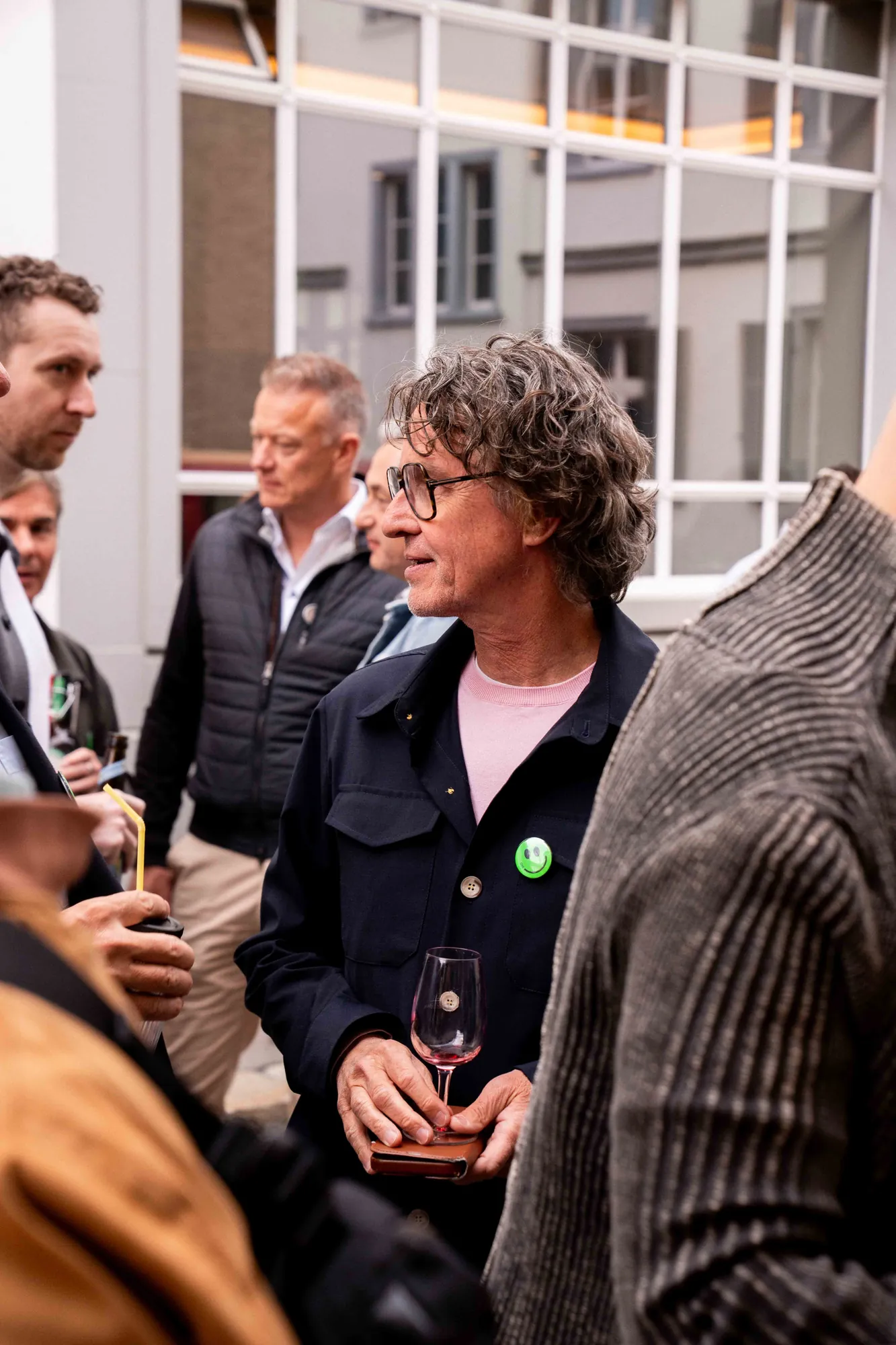 Gast mit lockigem grauem Haar, Brille und Weinglas hört in einer Menschengruppe vor einem Gebäude mit Sprossenfenstern zu.
