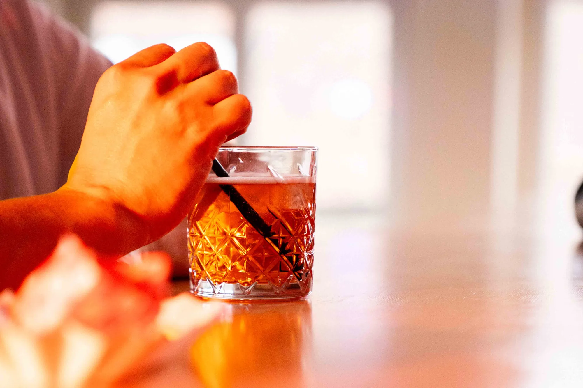 Hand rührt mit einem schwarzen Strohhalm einen bernsteinfarbenen Drink im geschliffenen Glas auf einem Holztresen.