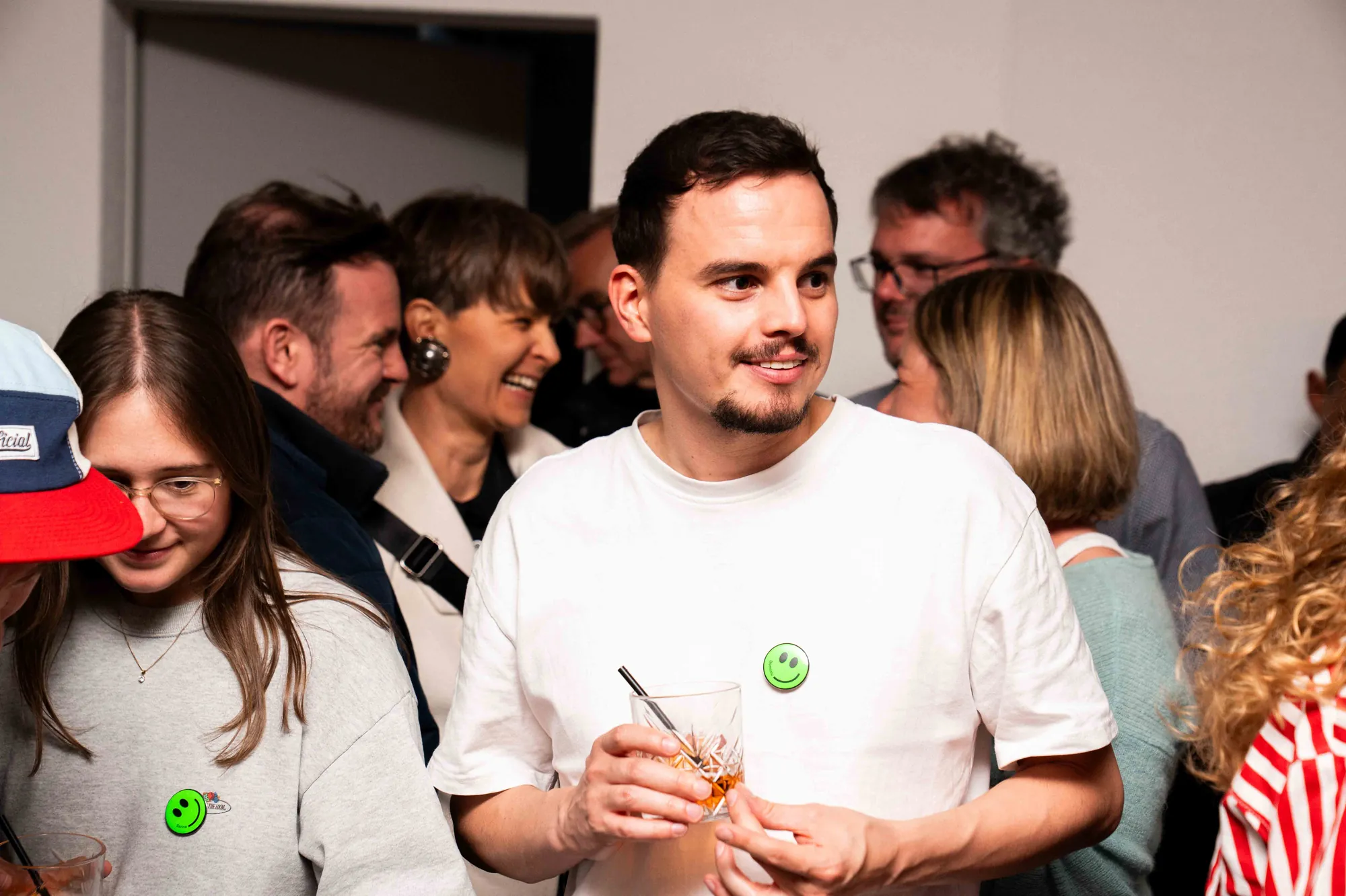 Mann im weissen T-Shirt mit Drink und grünem Smiley-Button lächelt inmitten einer dicht gedrängten Partygesellschaft.