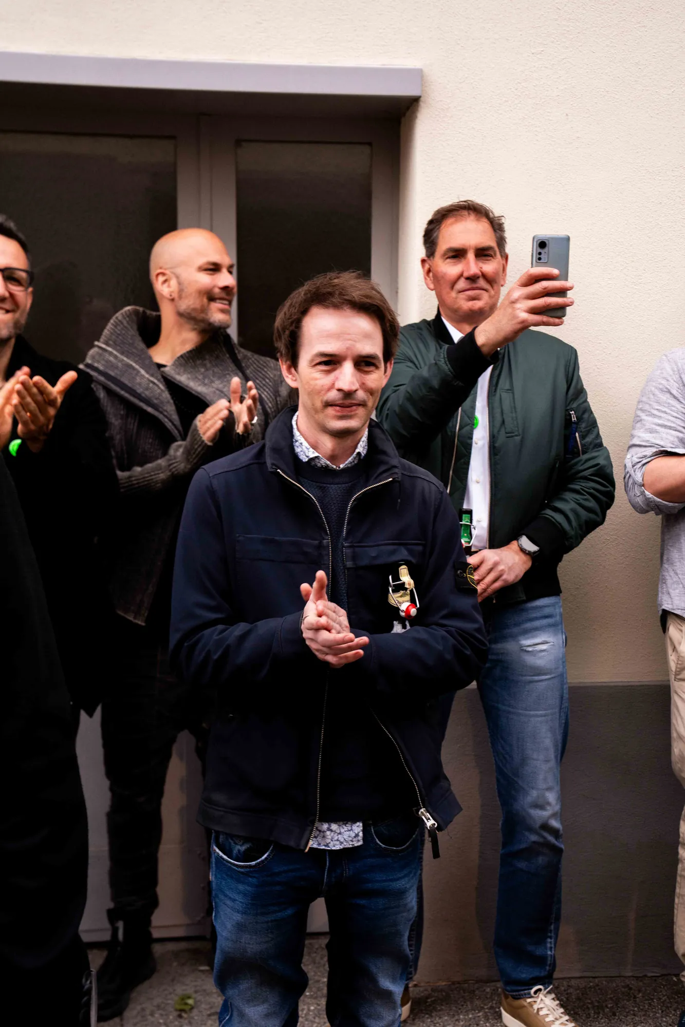 Mann in dunkler Jacke klatscht vor einer Hauswand, hinter ihm applaudieren weitere Gäste, einer hält das Smartphone zum Filmen hoch.