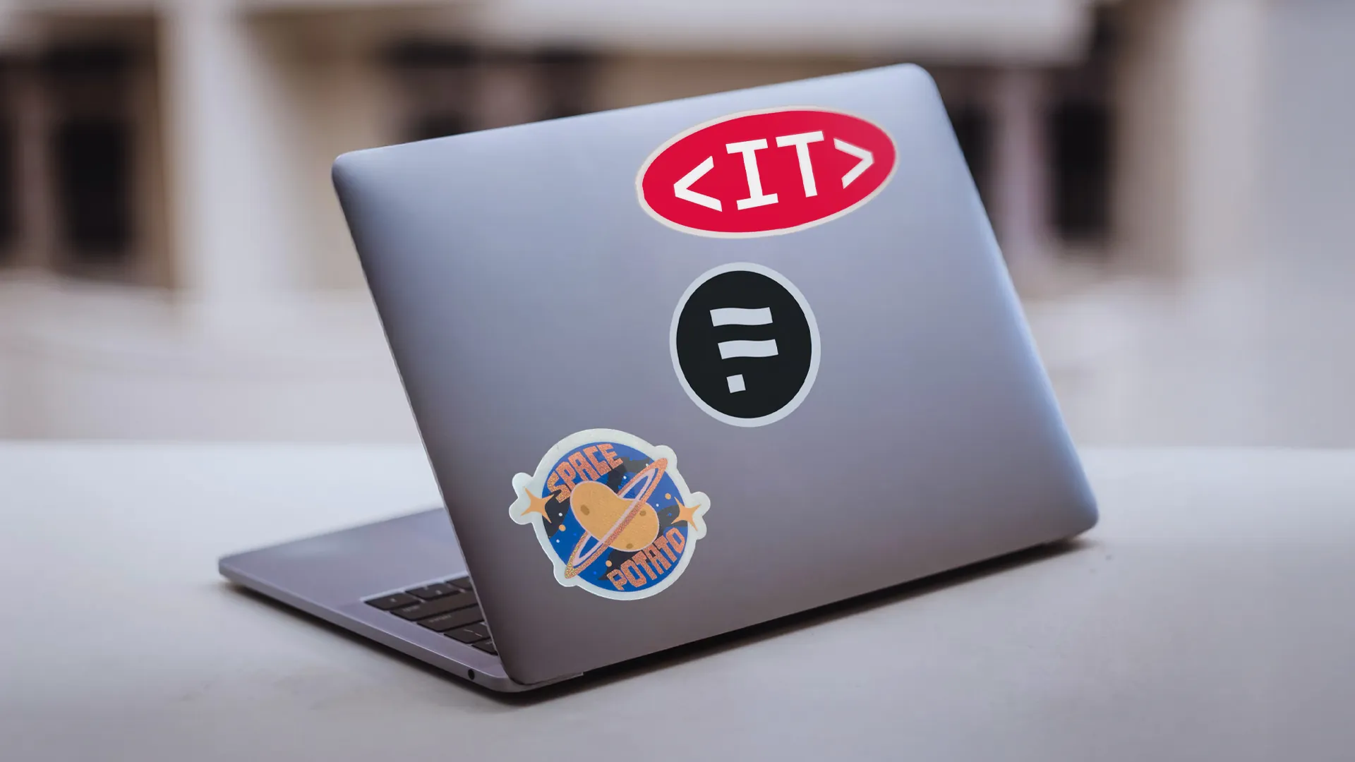 Aufgeklapptes silbernes Laptop von hinten mit Stickern, darunter ein rotes «<IT>»-Logo und ein schwarzes rundes Festland-Logo.