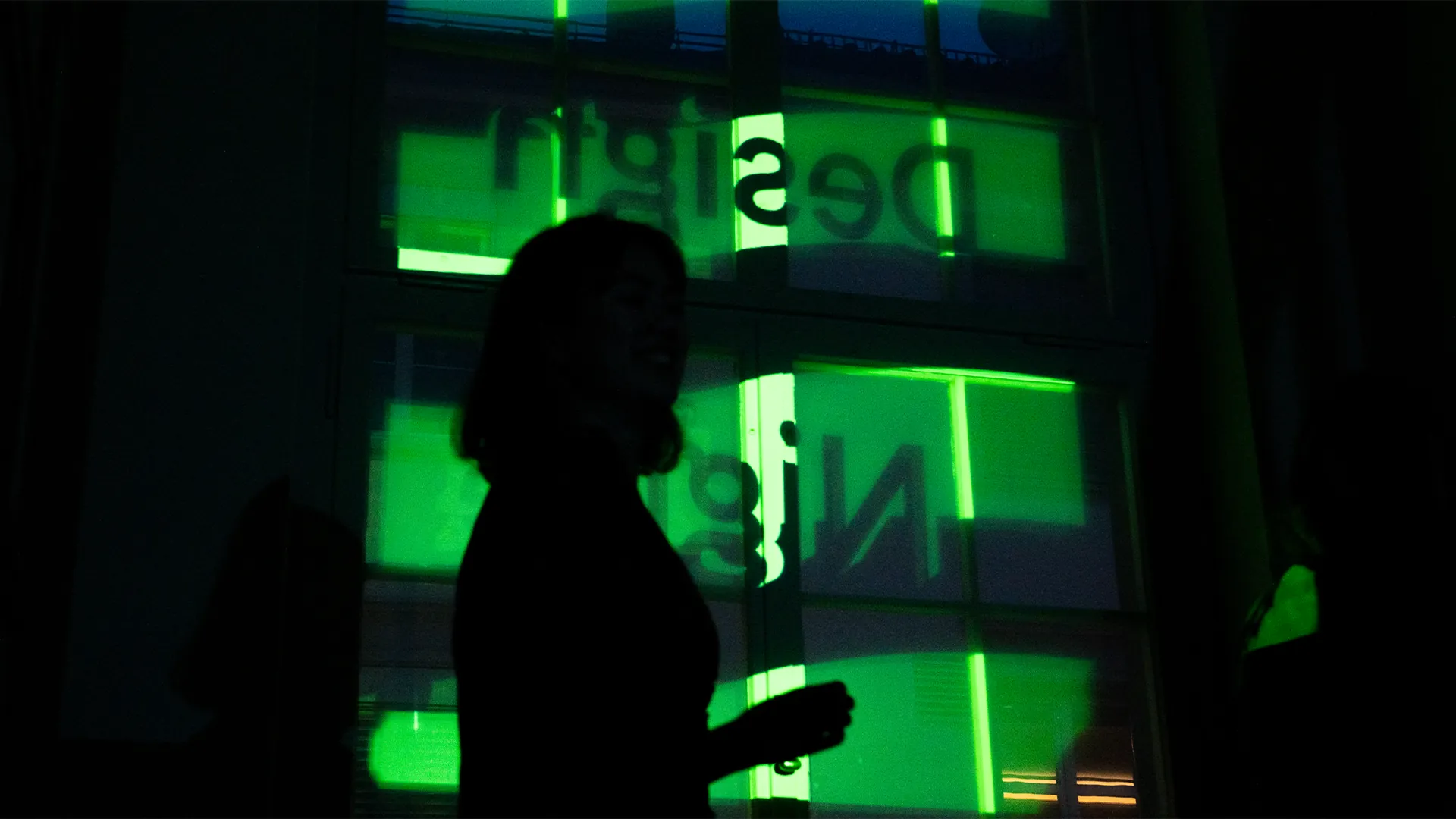 Silhouette einer Frau vor einem Fenster, auf das in leuchtendem Grün der Schriftzug «Design Night» projiziert ist.