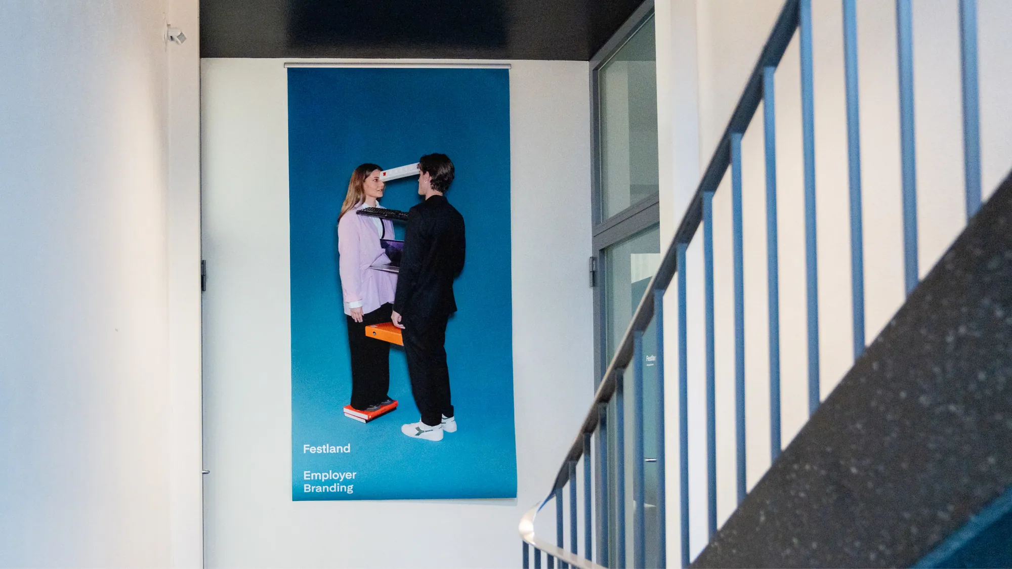 Treppenhaus mit grossem blauem Plakat «Festland Employer Branding», auf dem eine Frau und ein Mann in Schwarz einander gegenüberstehen.
