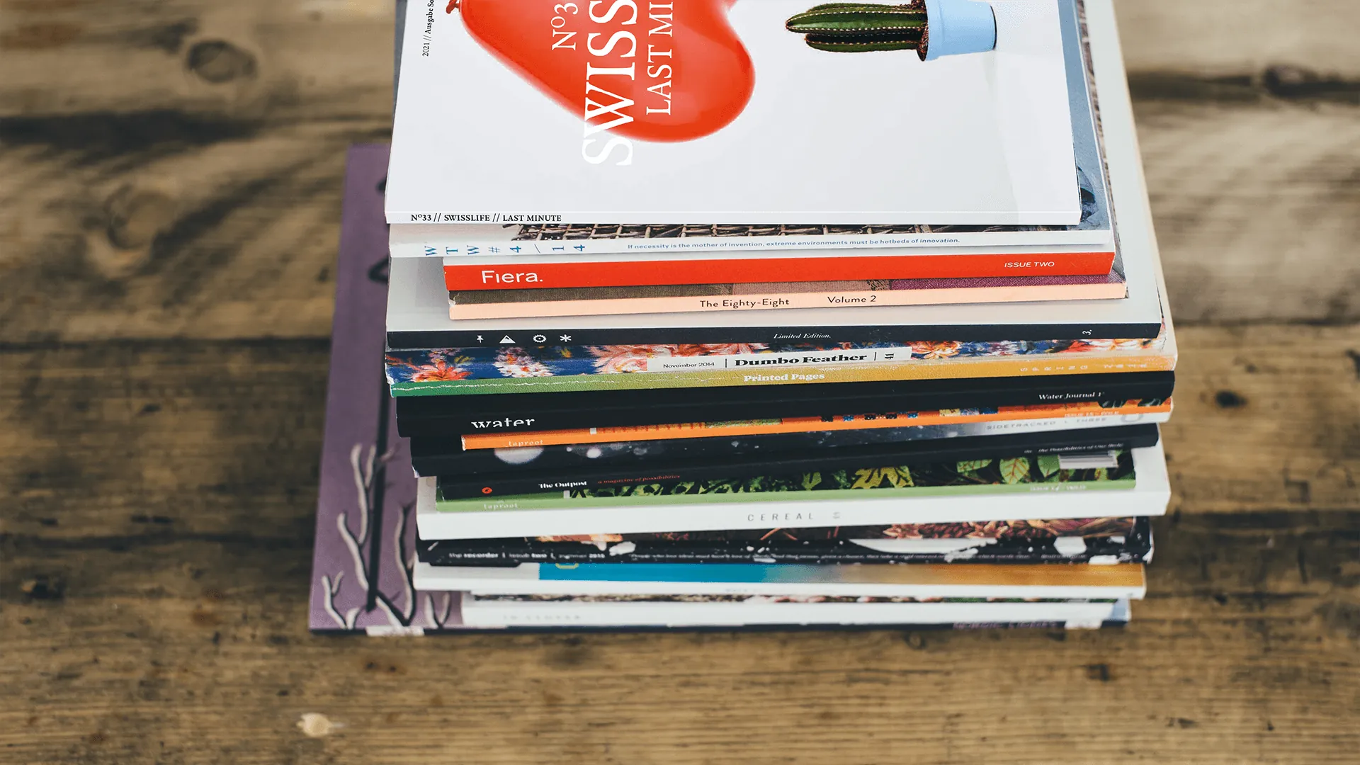 Stapel aus verschiedenen bunten Magazinen auf einem rustikalen Holztisch, zuoberst ein Heft mit rotem Herzmotiv und Kaktus.