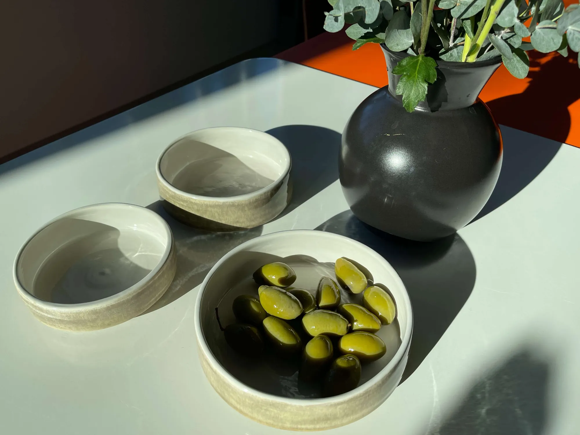 Drei fertig glasierte Apéroschalen im Sonnenlicht, eine mit grünen Oliven gefüllt, daneben eine schwarze Vase mit Eukalyptus.