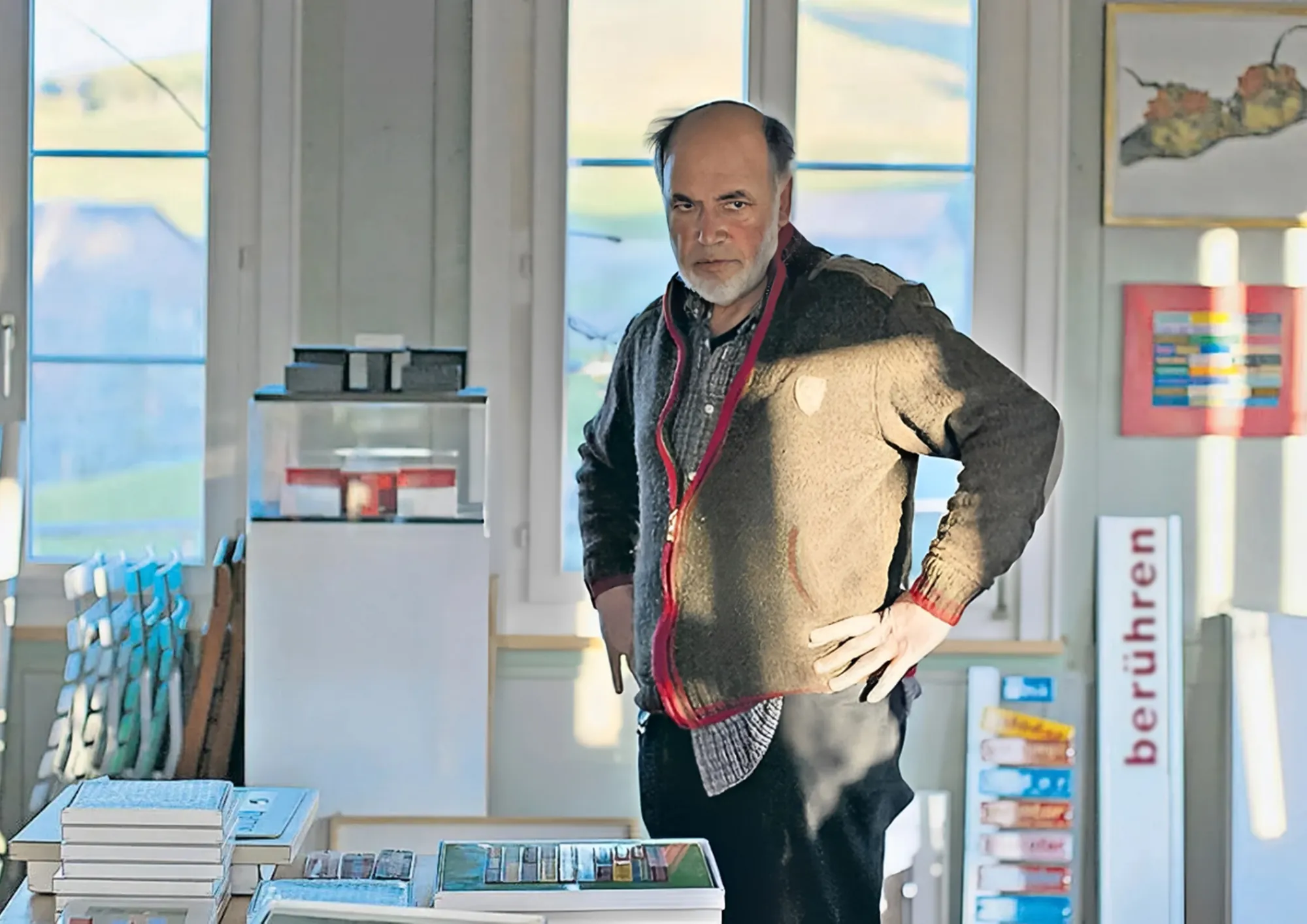 Mann mit grauem Bart steht in einem Raum mit Büchern, Kunstwerken und einem Schild mit dem Wort «berühren»