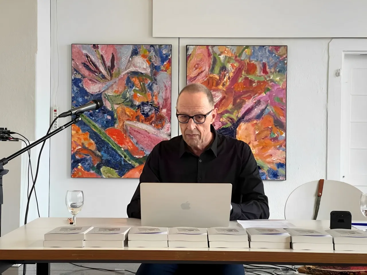 Lucas Roos liest an einem Tisch mit Laptop, Mikrofon und Stapeln seines Buches, dahinter zwei farbige Blumengemälde.