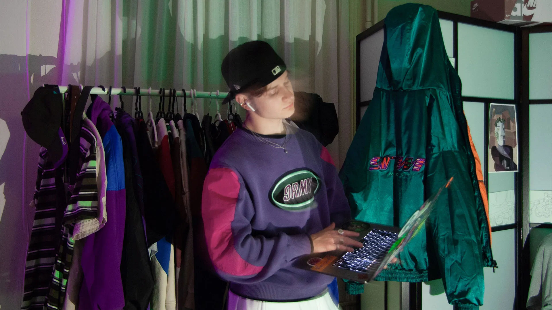 Valentin in violettem Retro-Pullover und Cap arbeitet am Laptop zwischen einer Kleiderstange mit bunten Jacken und einer grünen Windjacke.