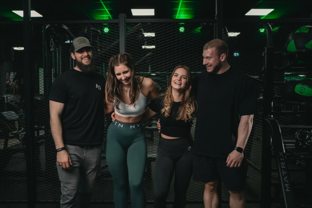 Four fit adults smiling and standing together inside a gym with fitness equipment in the background.