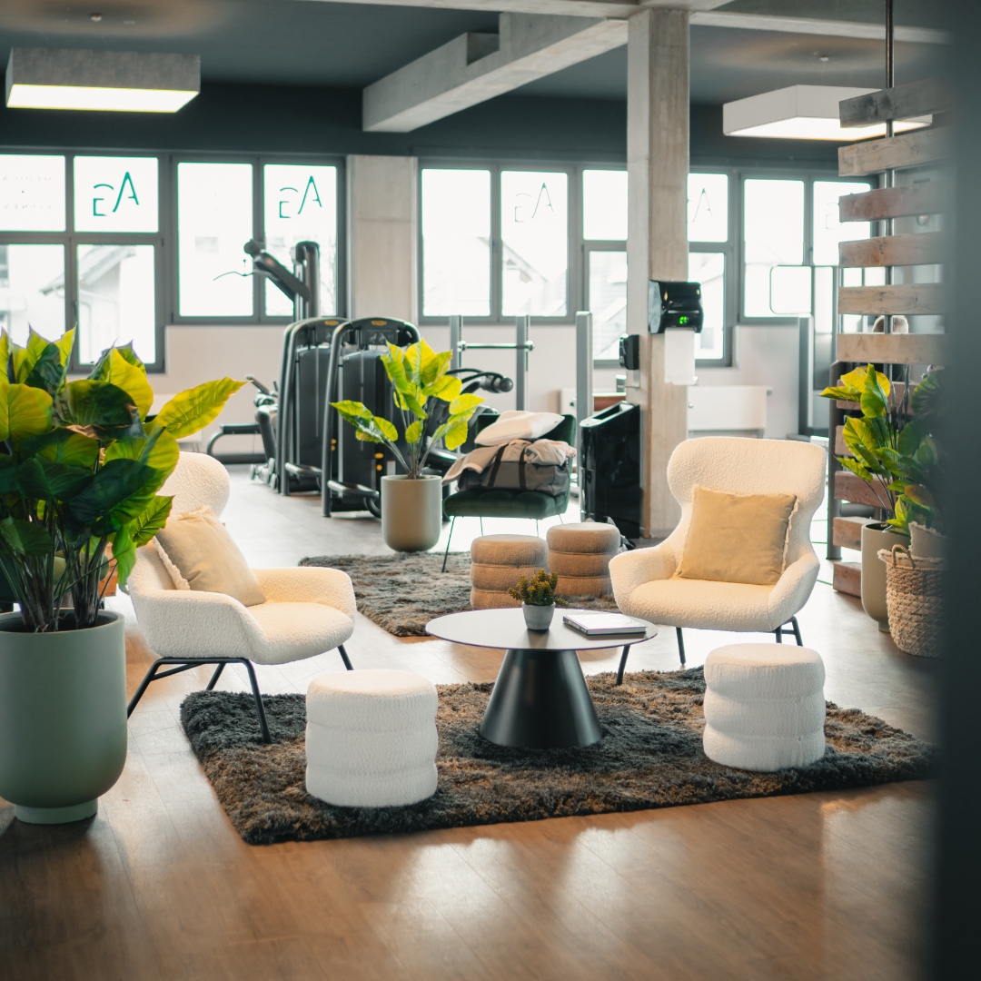 Modern fitness center interior with gym equipment and a comfortable lounge area featuring two armchairs, two ottomans, a round table, plants, and wood flooring.