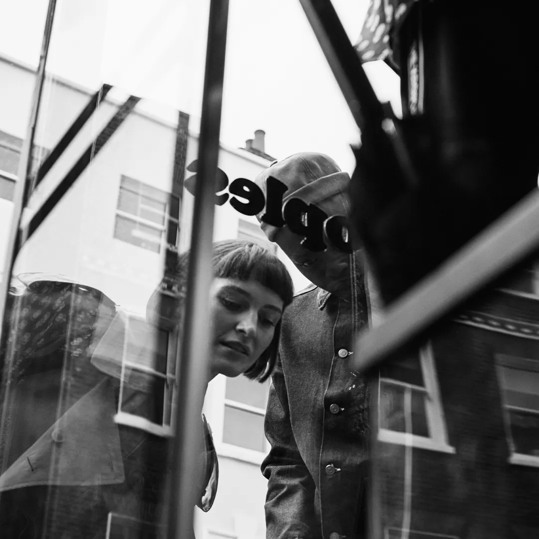 Black-and-white photo of two people looking into a store window with reflections of urban architecture.