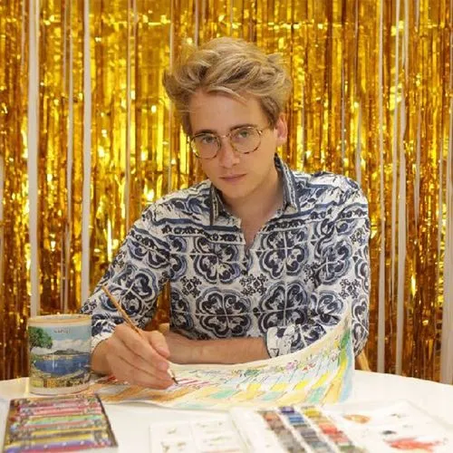 Artist wearing patterned shirt working on a colorful painting at a table, with a gold tinsel backdrop.
