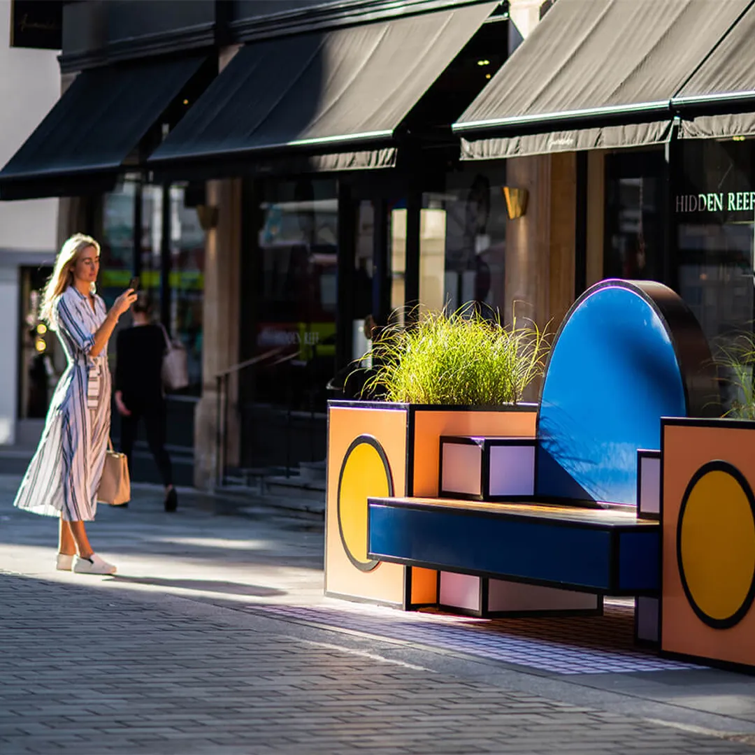 Woman in a striped dress walking past a vibrant geometric bench with plants on a sunny street.