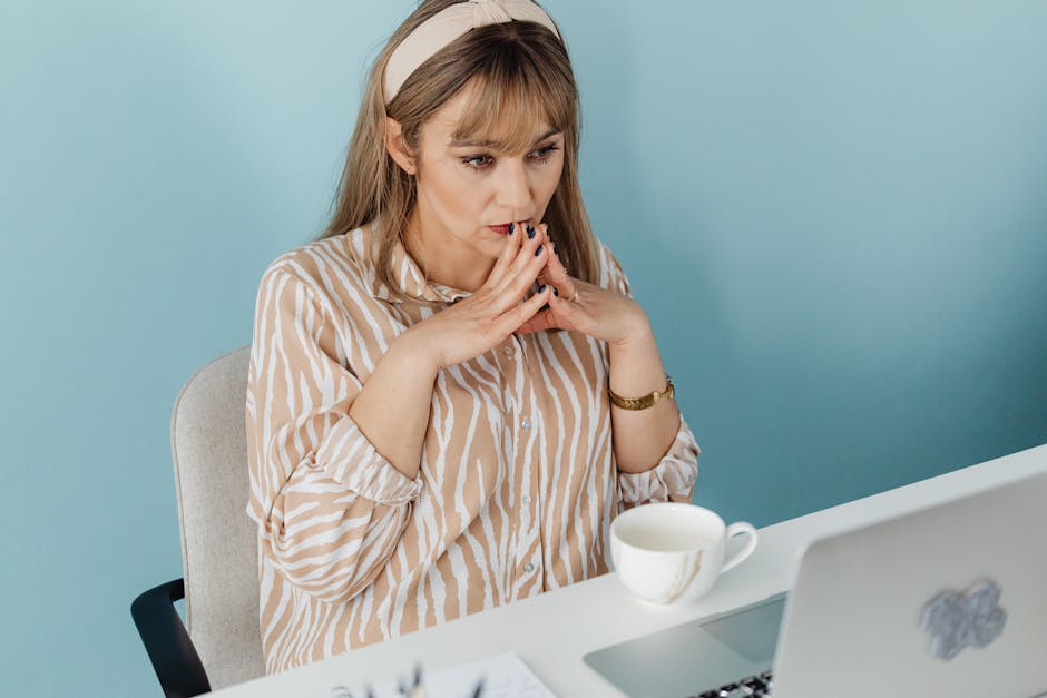 Thoughtful woman at desk with laptop and coffee, in a light blue office setting.