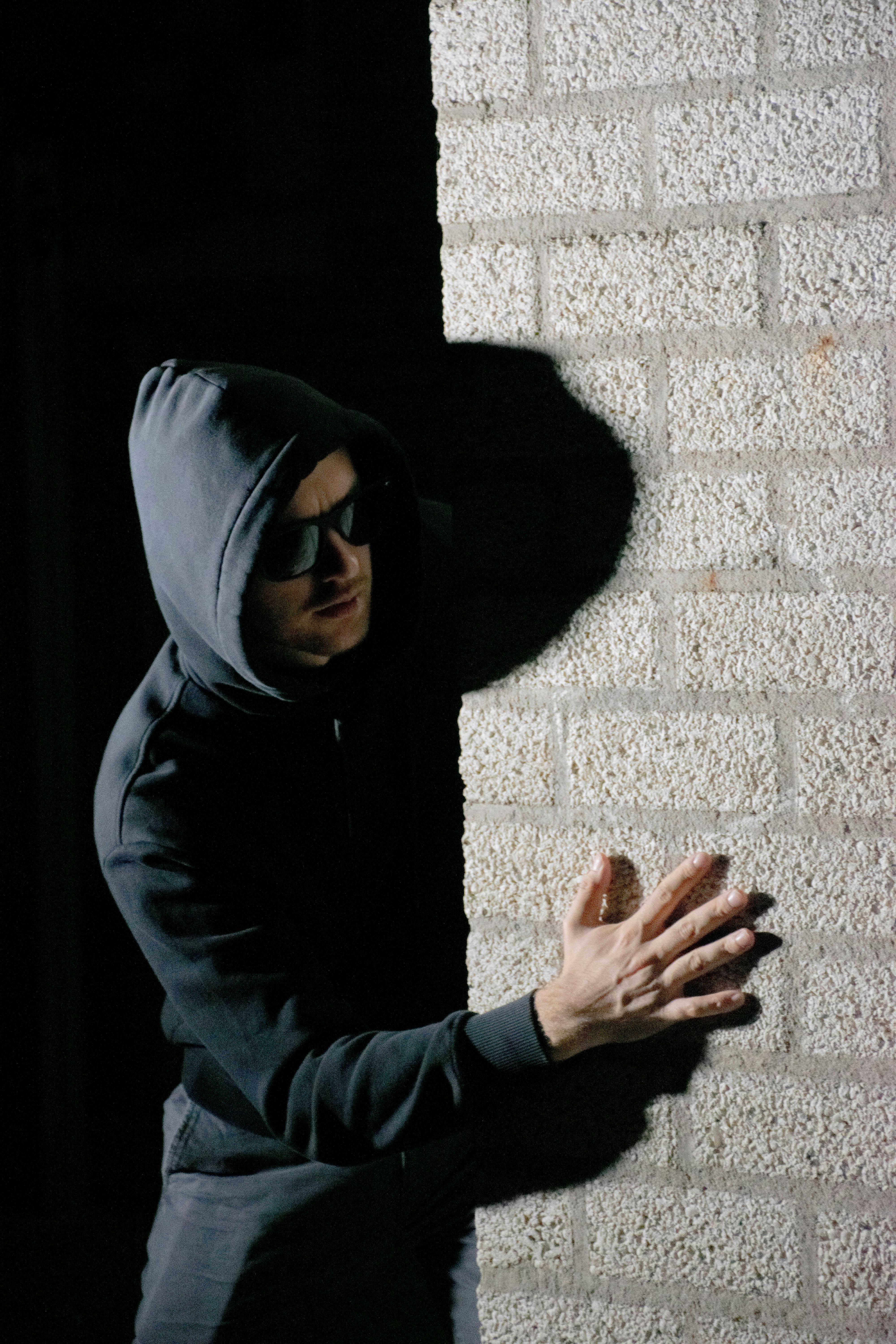 A man in a hoodie hides in the shadows against a brick wall, creating a mysterious mood.