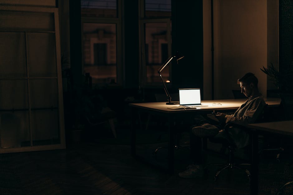 A man working late at night in a dimly lit office, with a bright laptop screen.