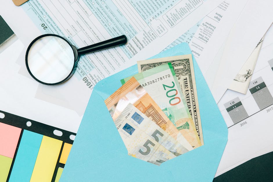 Close-up of tax documents and mixed currency in a blue envelope with a magnifying glass, perfect for financial analysis concepts.
