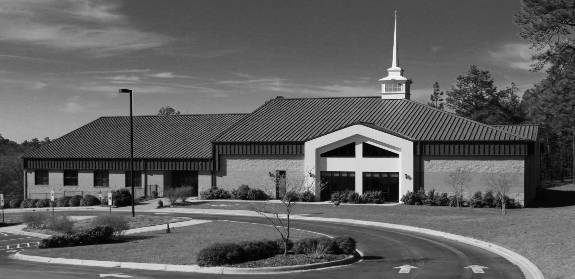 Black-and-white exterior photo of a single-story stone and stucco church building with a steep metal roof, tall white steeple topped with a cross, and a glass entryway framed by a large cross-shaped window, located in Southern Pines, North Carolina.