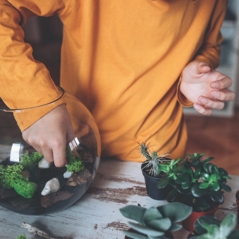 Child in an orange long sleeve shirt assembling a terrarium inside a glass sphere.