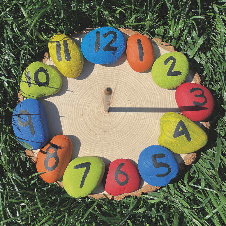 Homemade wooden sundial with colorful rock hours and pencil pointer on grass.