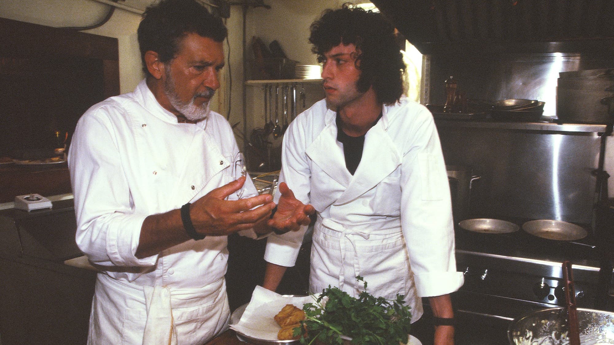 An older chef in whites talks a young cook through a dish in a restaurant kitchen, in a still from Tony.