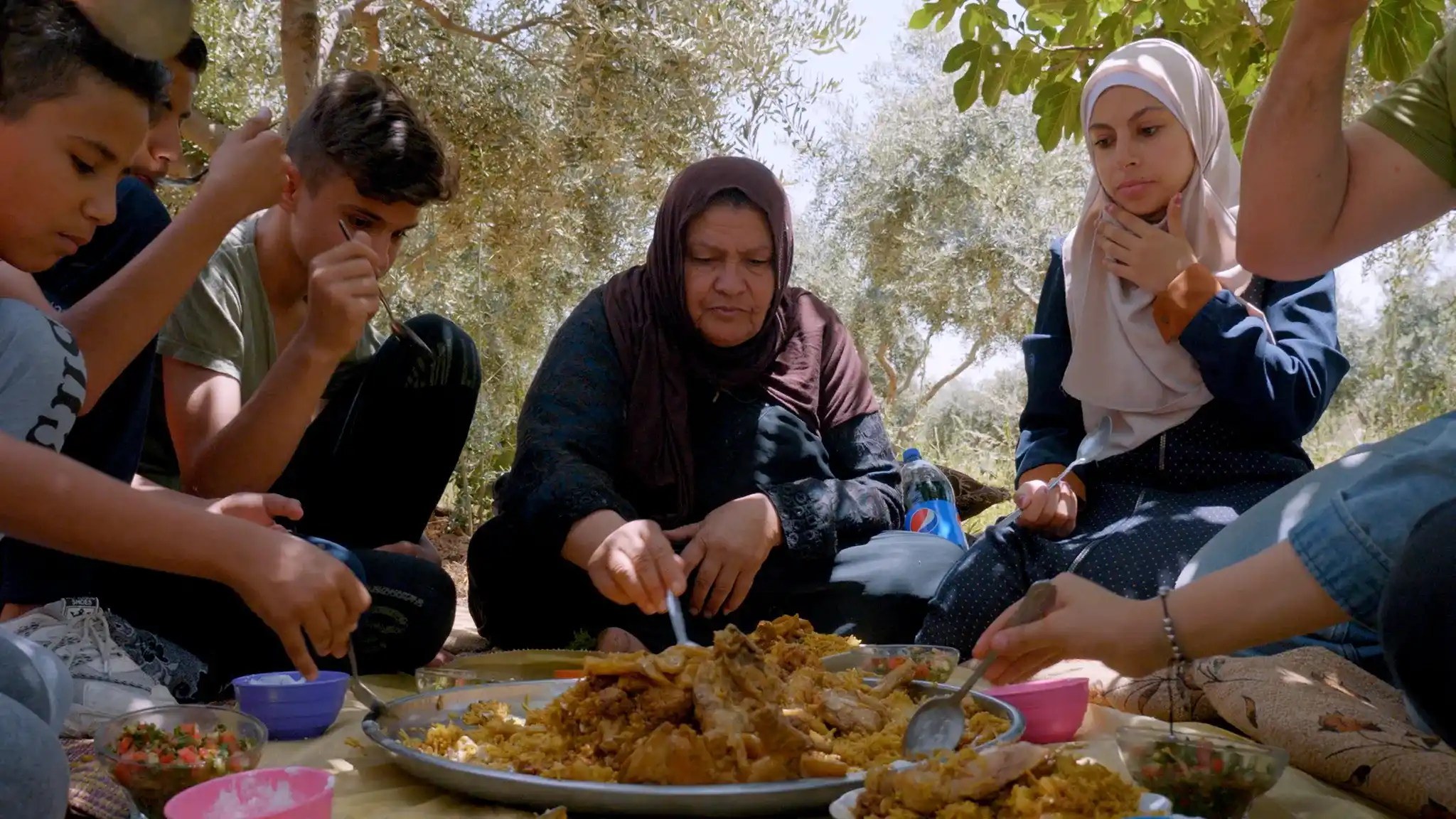 A family shares a large platter of maqloobeh outdoors under olive trees in a still from the documentary Aisha's Story.