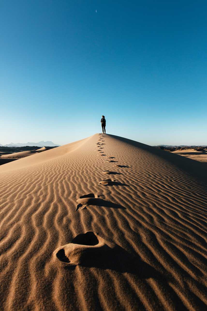Person standing on top of a sand dune with a clear blue sky above and a trail of footprints leading to them in the desert.