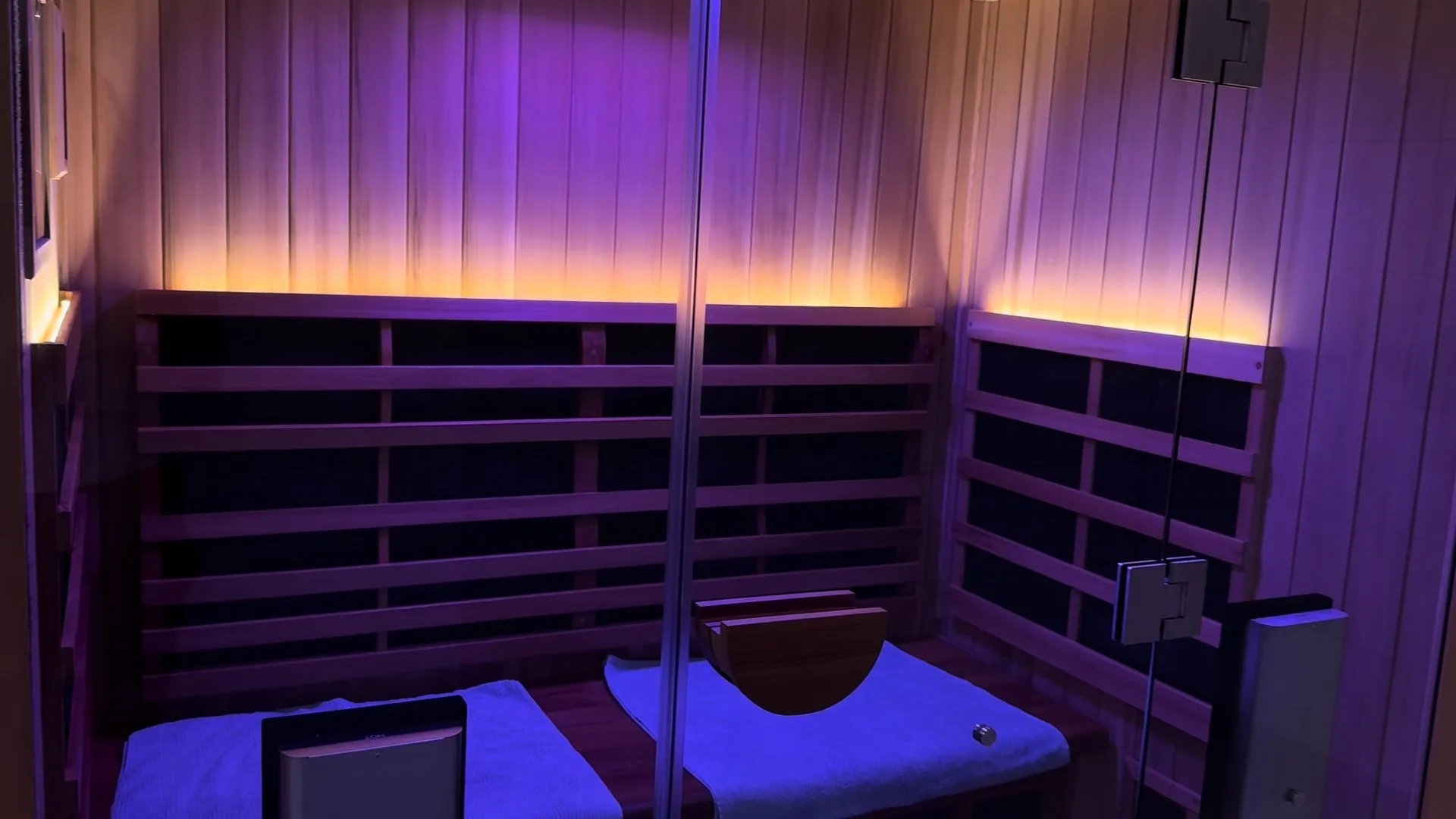 Interior of a wooden sauna room with two benches covered by blue towels and soft purple lighting.