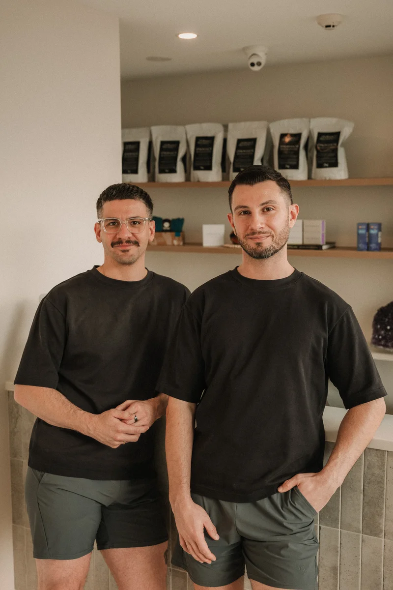 Two men in black shirts and dark shorts standing indoors with shelves of products behind them.