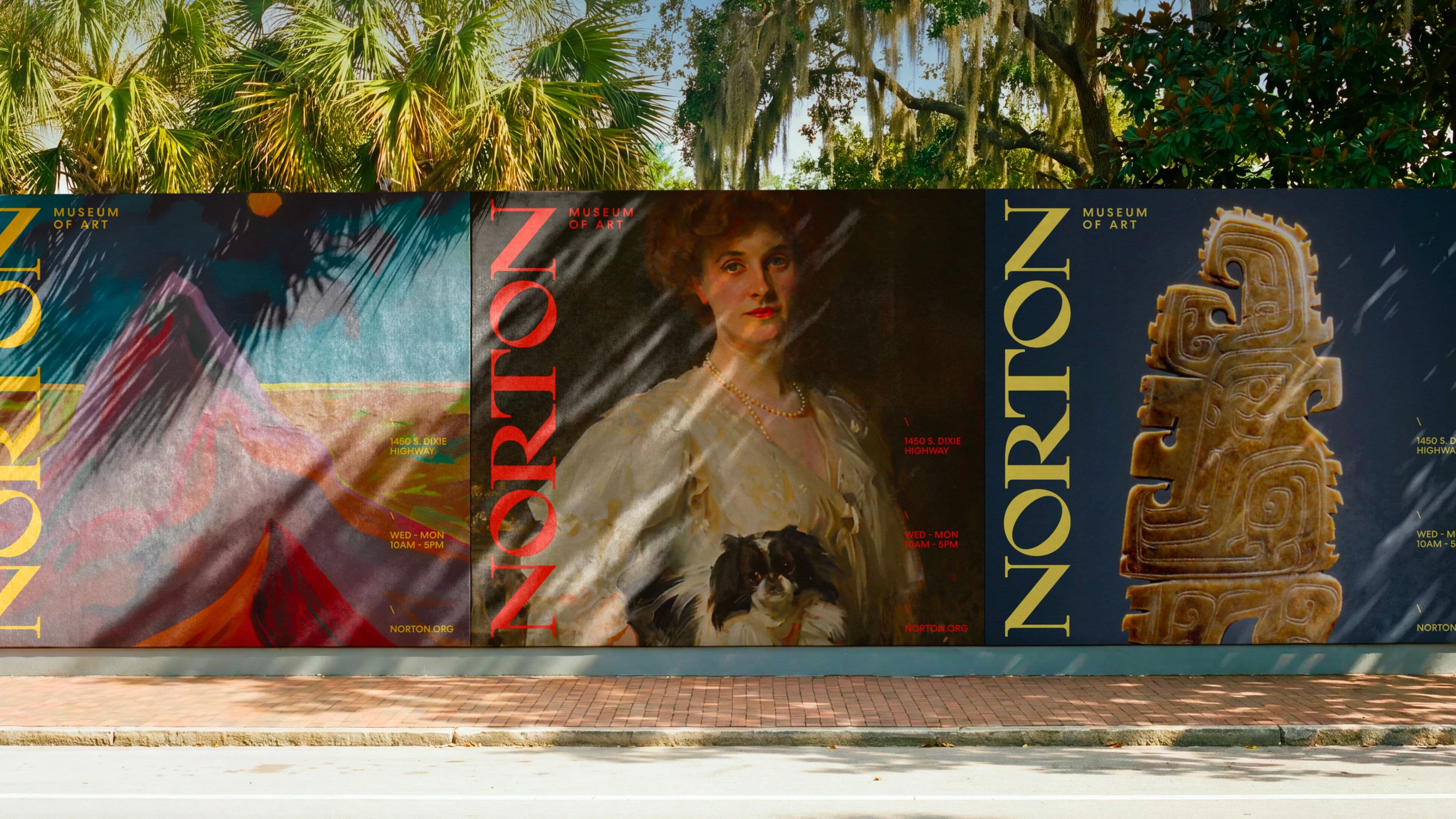 A street-facing hoarding wall featuring a series of Norton Museum of Art branded panels, each showcasing a different work from the collection — including a colorful abstract painting, a classical portrait of a woman holding a small dog, and an ancient carved artifact. Panels include the museum's address, hours, and website.