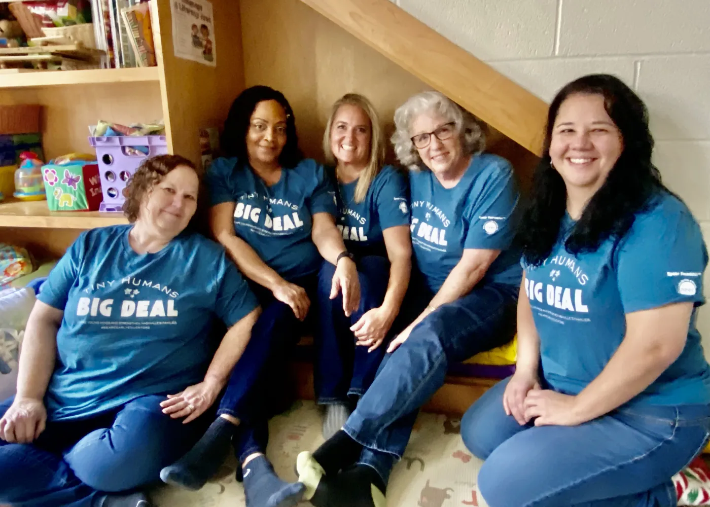 Five women seated indoors wearing matching blue T-shirts that say 'Tiny Humans Big Deal,' smiling and posing together.