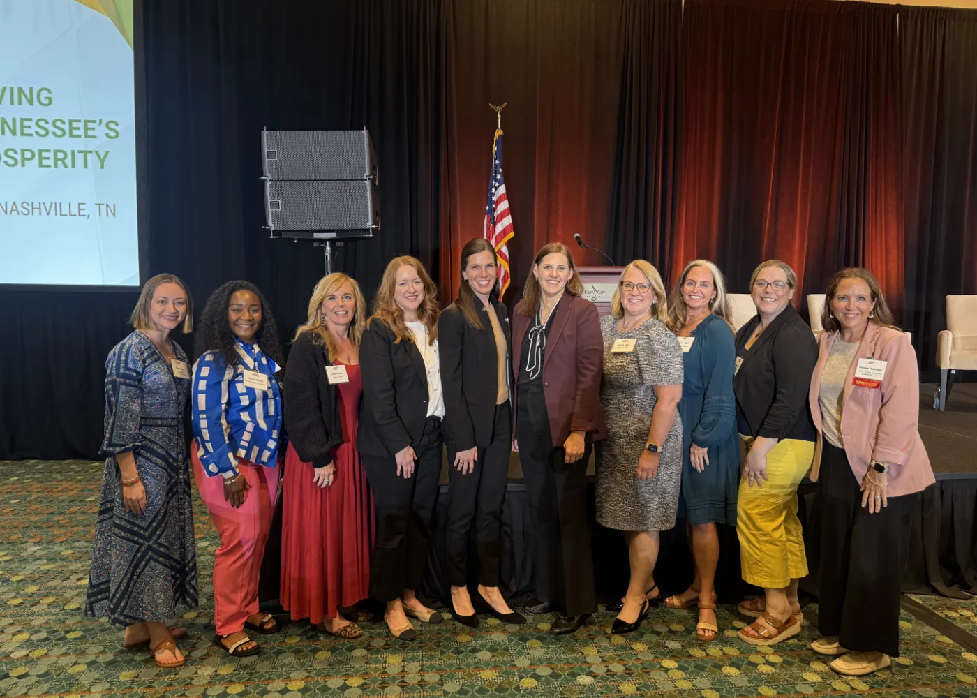 Group of ten professionally dressed women standing in a line smiling at a conference with an American flag and stage in the background.