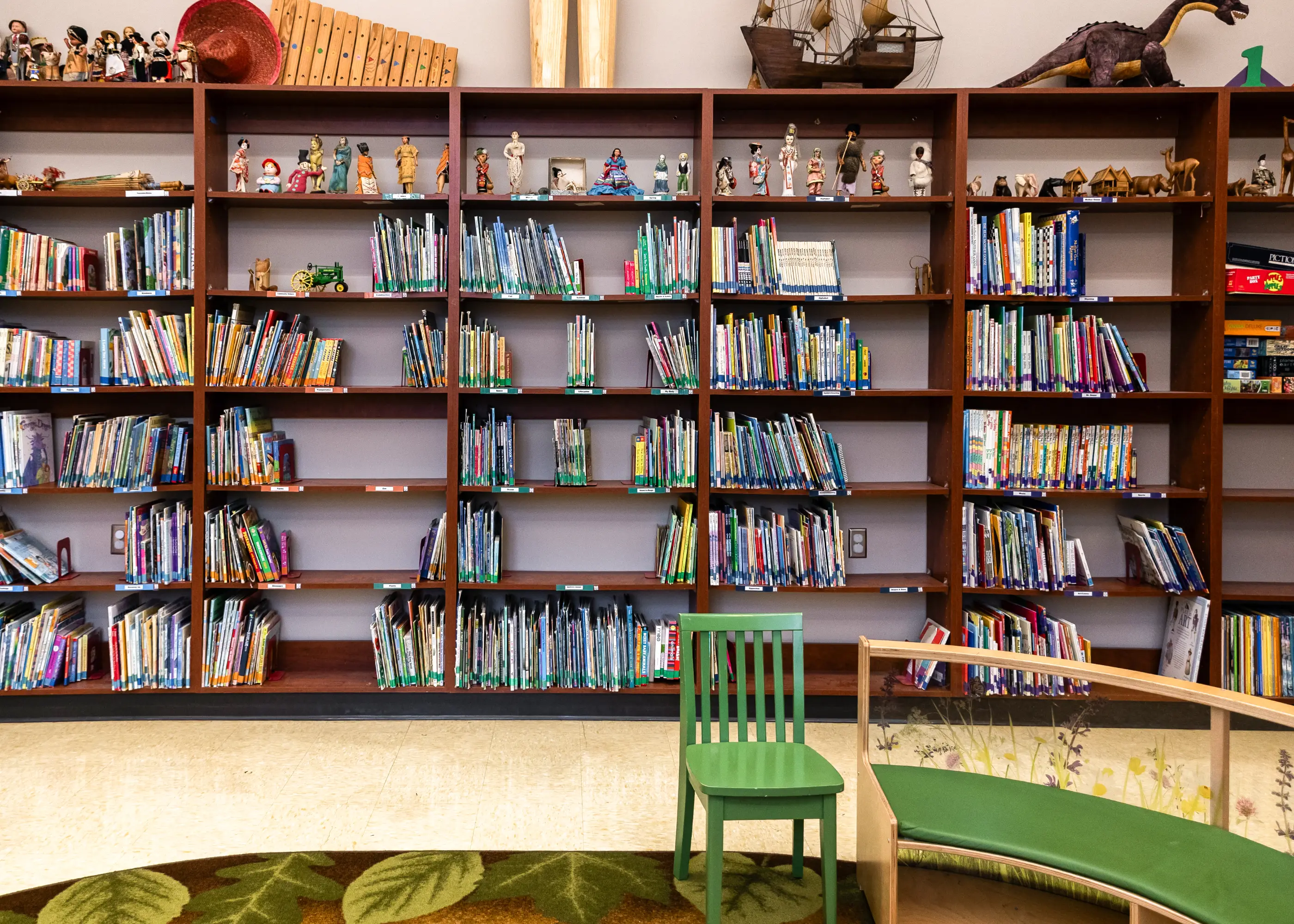 Children’s library with wooden bookshelves filled with books and cultural figurines, a green chair, and a curved wooden bench with green cushions.