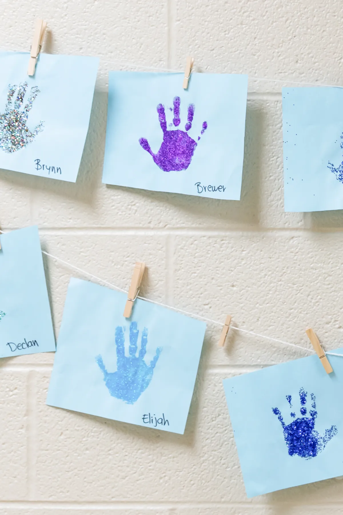Blue papers with colorful glitter handprints and children's names pinned on a wall with clothespins.