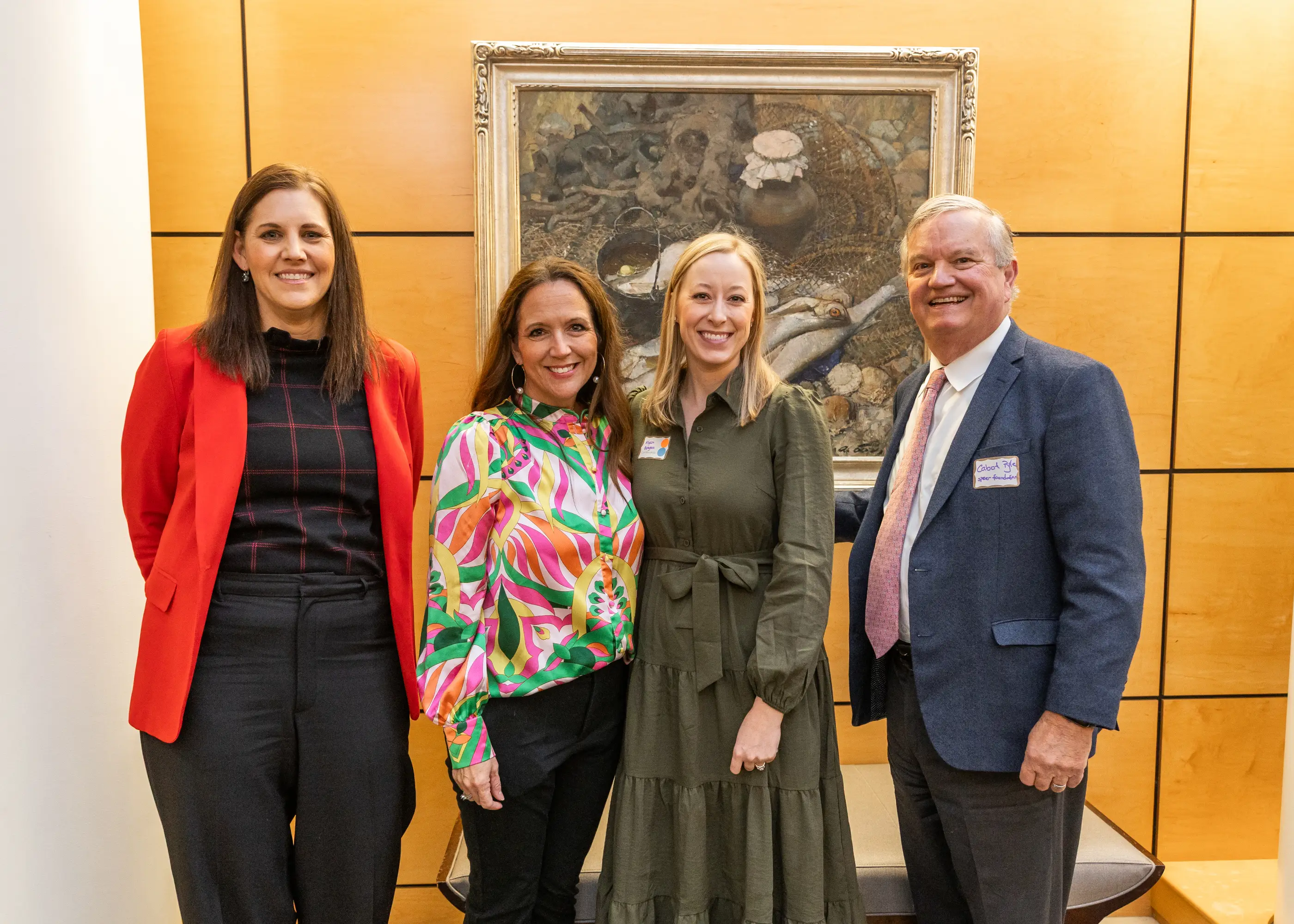 Four people smiling and posing indoors in front of a framed painting on a wood-paneled wall.
