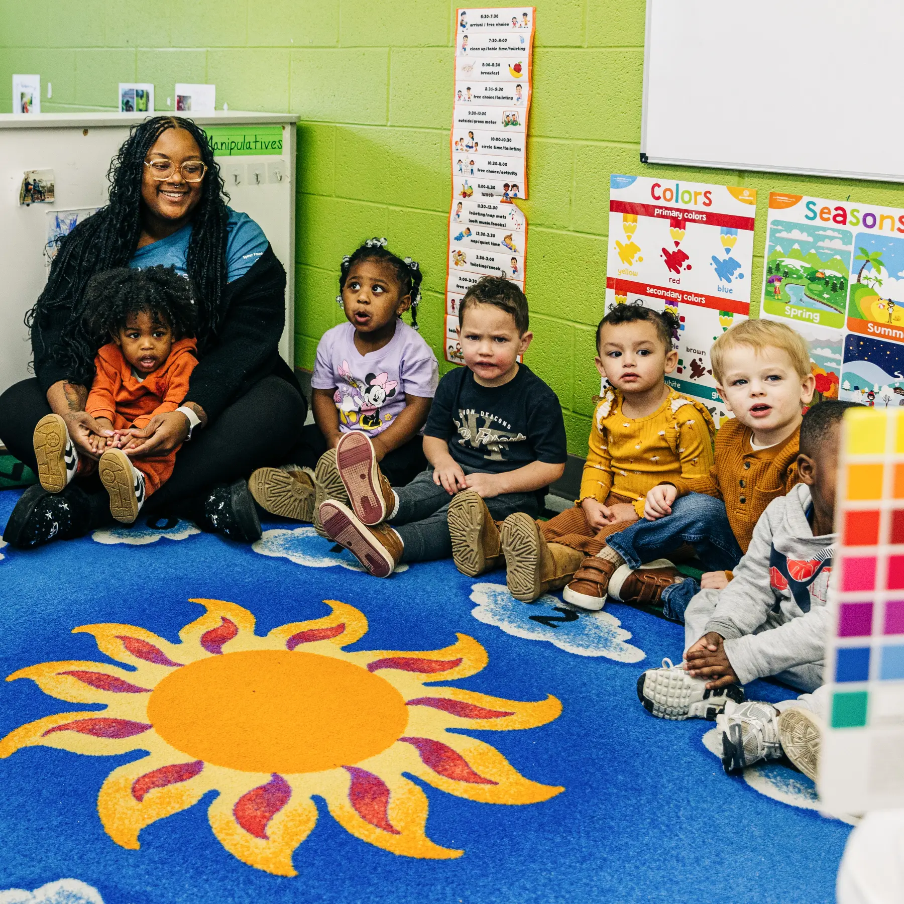 Teacher sitting on a colorful carpet with six young children in a classroom with educational posters on seasons and colors.