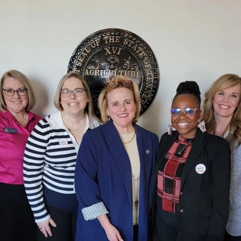 Five women standing together smiling in front of the State of Tennessee Agriculture seal on a wall.