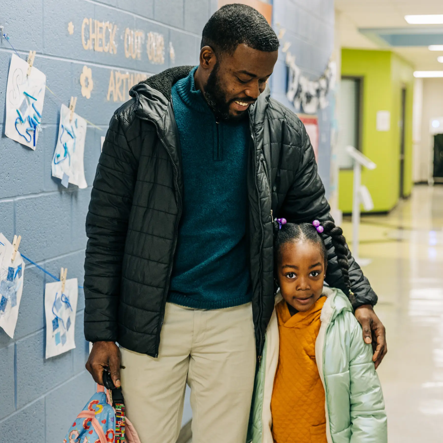 Smiling parent with arm around his young daughter standing in a school hallway with children's artwork on the wall.