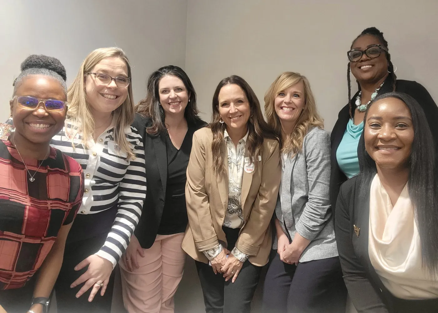 Group of seven diverse women smiling and posing together against a plain wall.