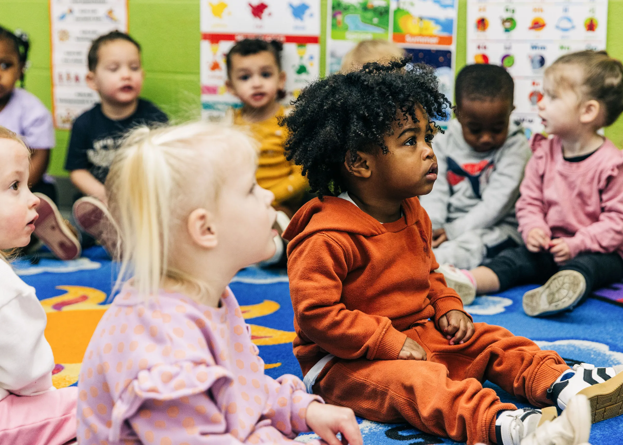 A group of young children sitting on a colorful classroom rug, attentively looking forward during a learning activity.