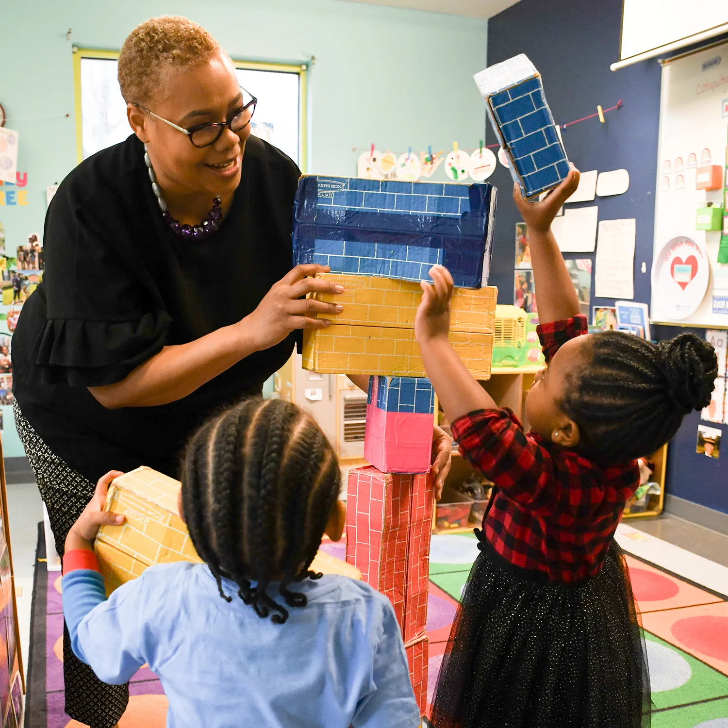 Teacher and two children building a tall tower with colorful cardboard blocks in a classroom.