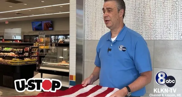 A person in a blue polo holds a folded American flag inside a convenience store. Nearby are shelves with food items. Logos for U-Stop and KLKN-TV are visible. The mood is respectful.